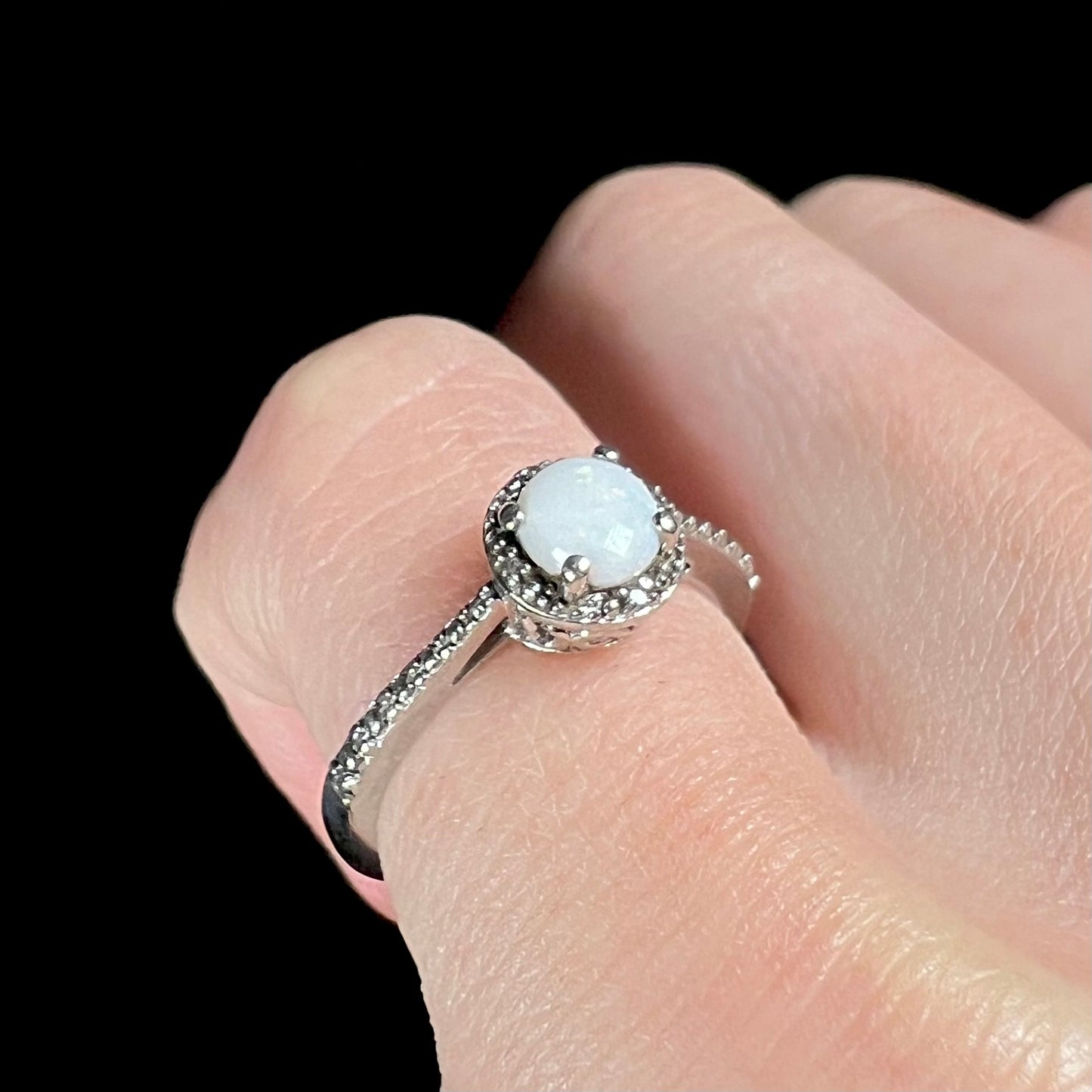 Close-up angled view of a sterling silver white opal and diamond ring with a halo design, worn on a pinky finger.