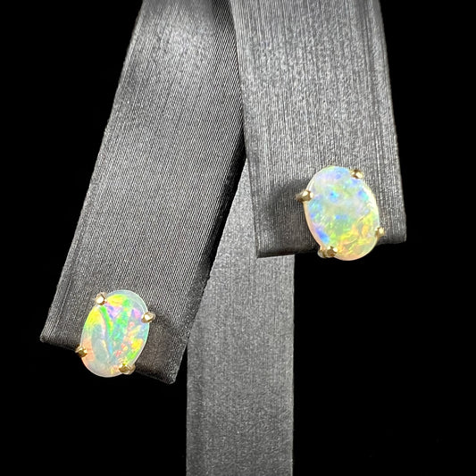 A pair of white opal stud earrings in 14k gold, displayed on a black background. The opals exhibit mostly yellow fire.