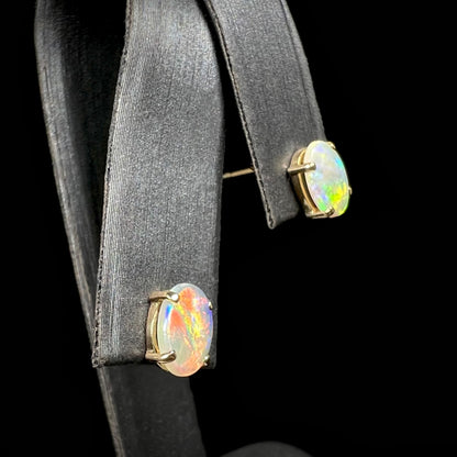 A close-up of white opal earrings in 14k gold, featuring a broad red flash in one opal and green fire in the other.