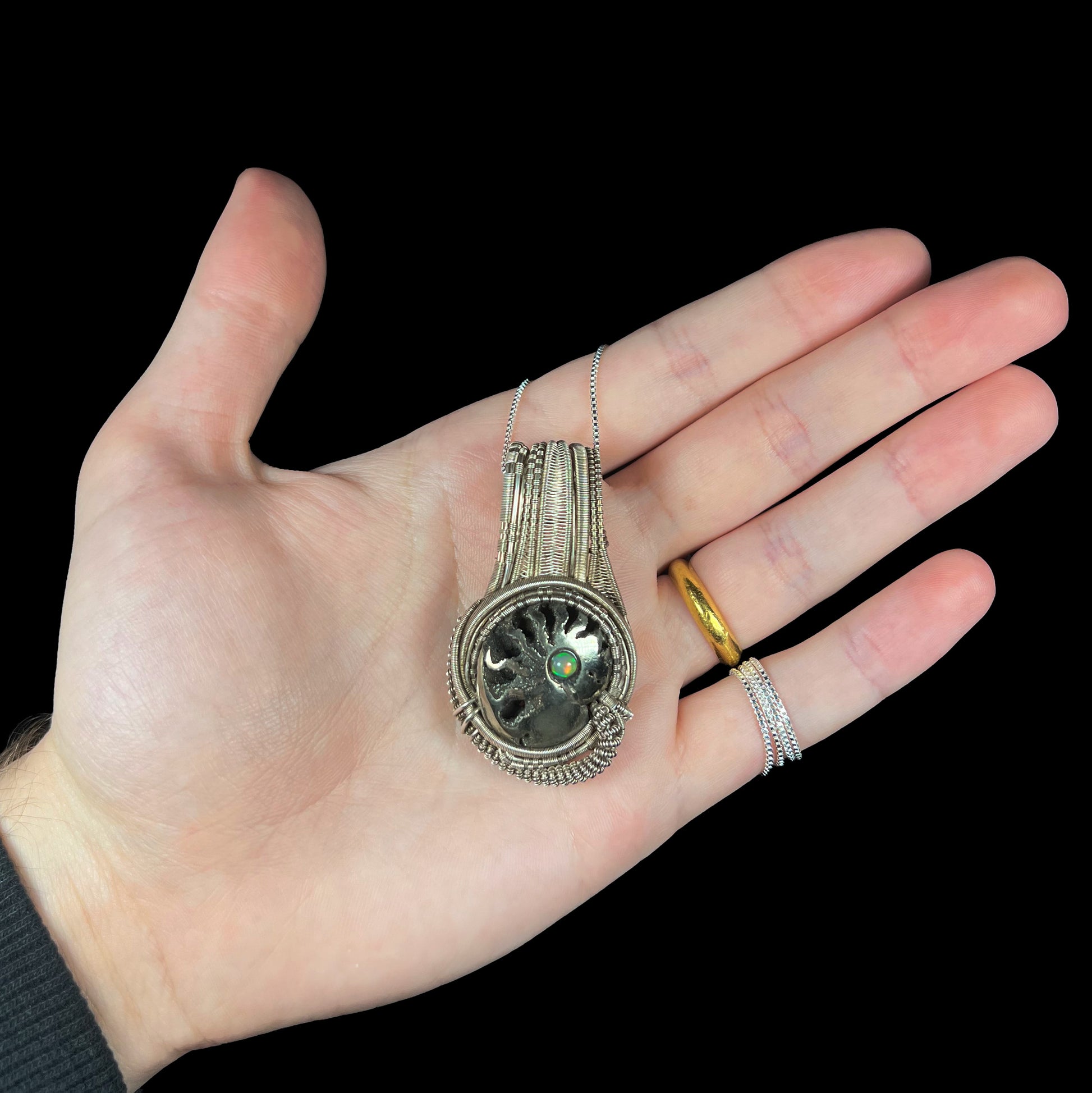 Hand holding a man's wire wrapped ammonite shell and opal-accented pendant on silver box chain.