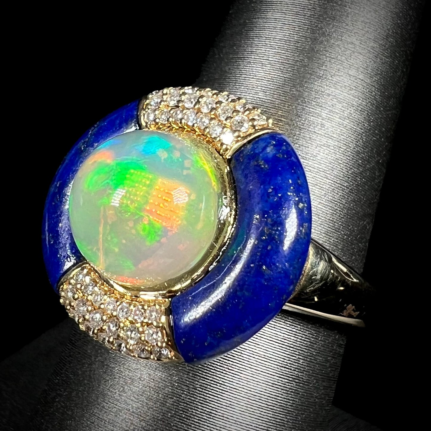 Angled close-up of a women's 14k gold Ethiopian fire opal, carved blue lapis lazuli, and diamond pave ring, shown on ring display.