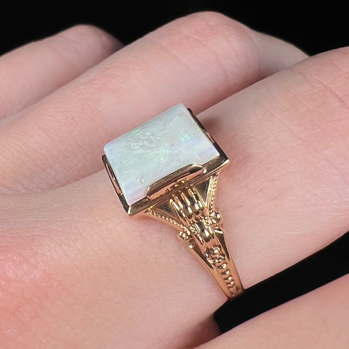 Close-up of finger wearing a women's Art Deco opal solitaire ring, the ring showing its ornate 10k yellow gold shoulder and shank