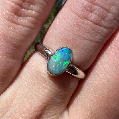 Close-up of a women's Australian opal solitaire ring in sterling silver, worn on a finger in natural sunlight.  Green and blue colors are seen in the opal.