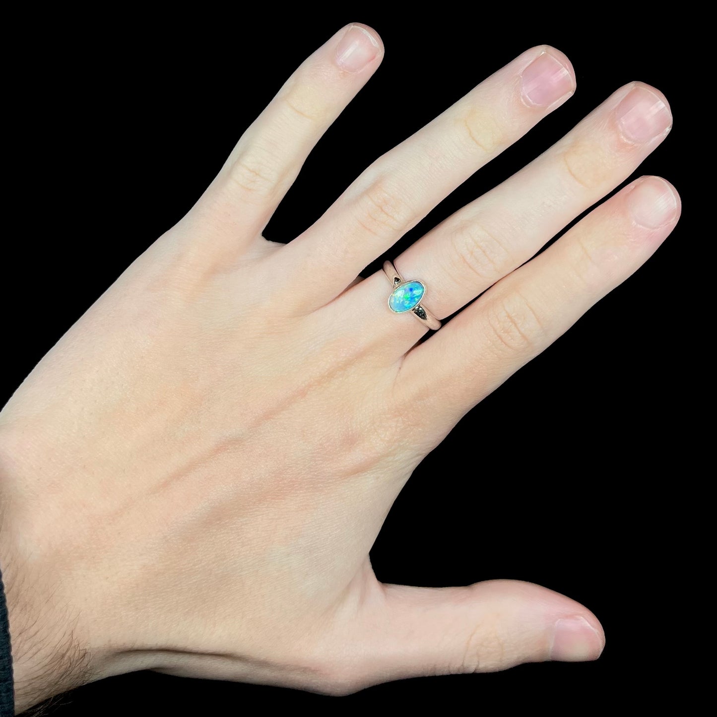 Hand wearing a women's bezel set sterling silver opal solitaire ring against black background.