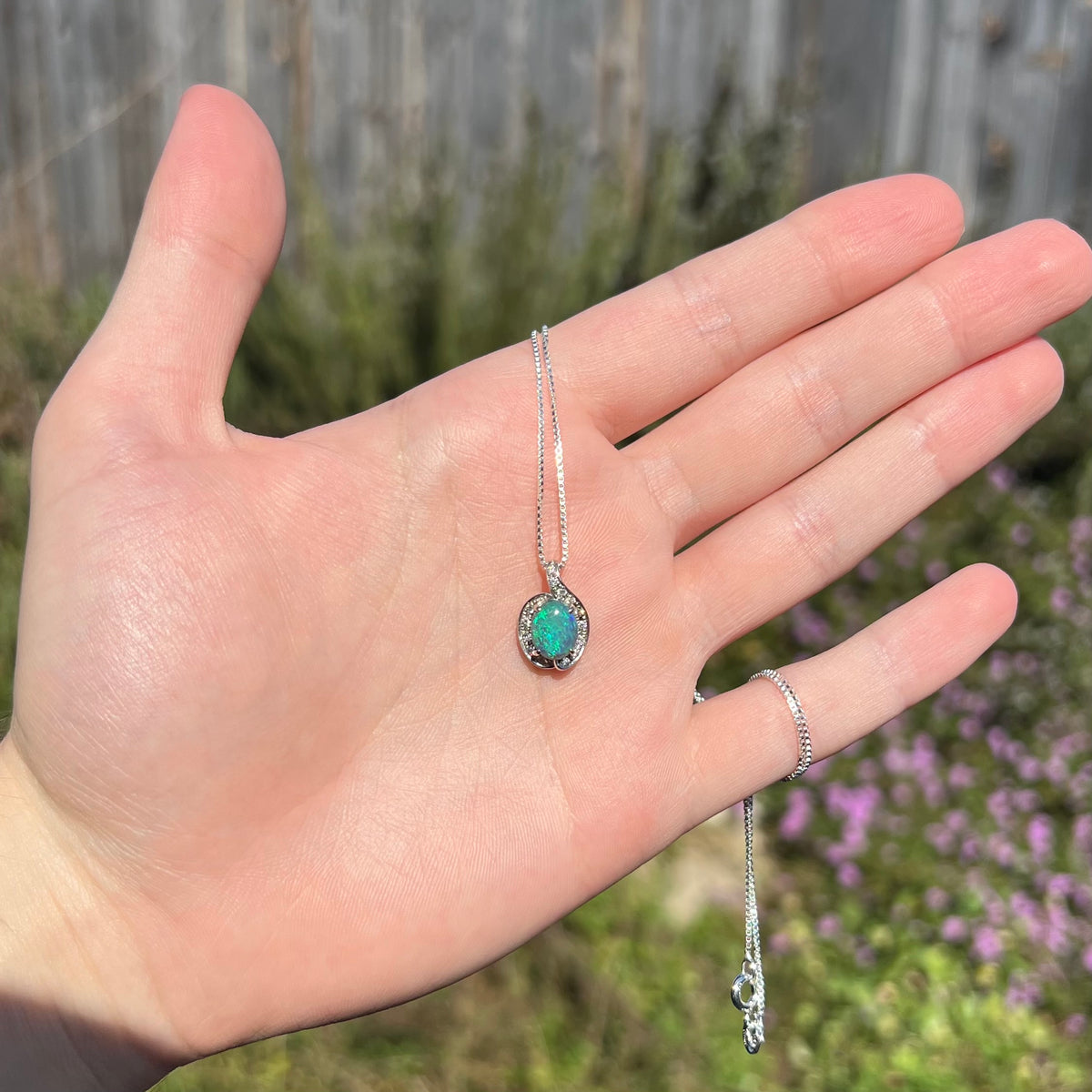 Sunlit hand holding women's black opal and diamond pendant against outdoor background.  The opal has a green flash of color.