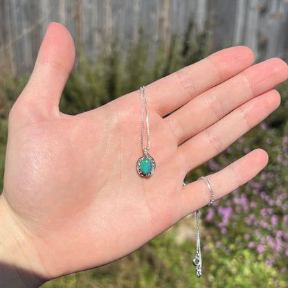 Sunlit hand holding women's black opal and diamond pendant against outdoor background.  The opal has a green flash of color.