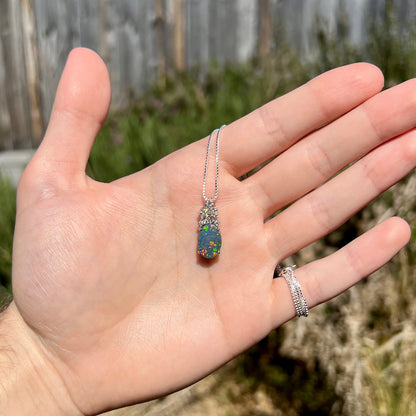 Hand holding women's black opal pineapple necklace in sunlight, red and green play-of-color seen in opal.
