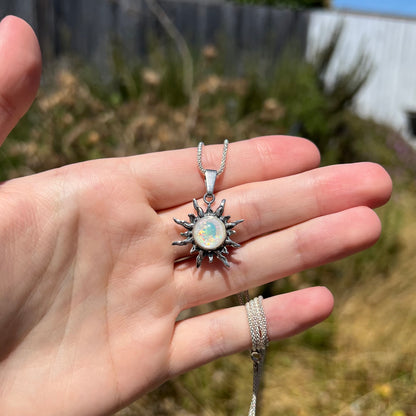 Hand holding a women's Boho-style sunburst pendant in sterling silver set with a round imitation opal in outdoor sunlight.