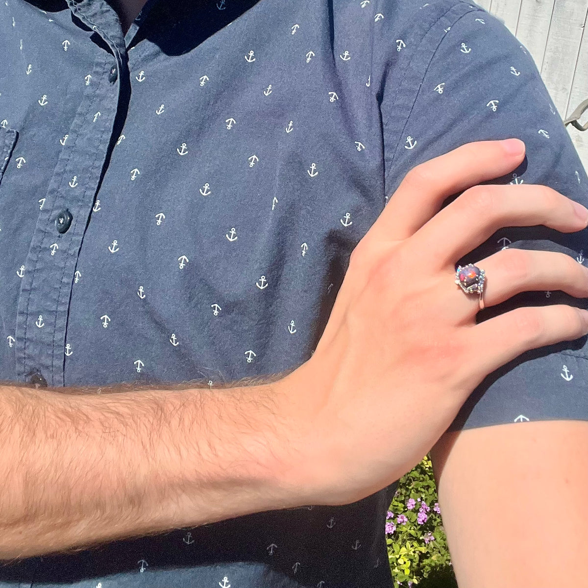 Close-up of person's hand resting on opposing shoulder in sunlight wearing a women's dark opal and diamond ring.
