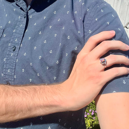 Close-up of person's hand resting on opposing shoulder in sunlight wearing a women's dark opal and diamond ring.