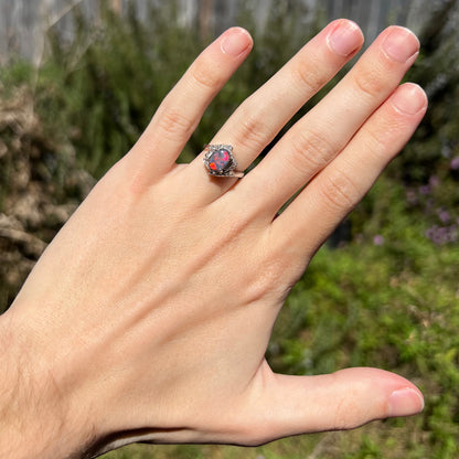 Hand wearing an Australian dark opal and diamond women's ring with vintage Japanese platinum design under natural sunlight.  The opal has bright red and magenta play-of-color.