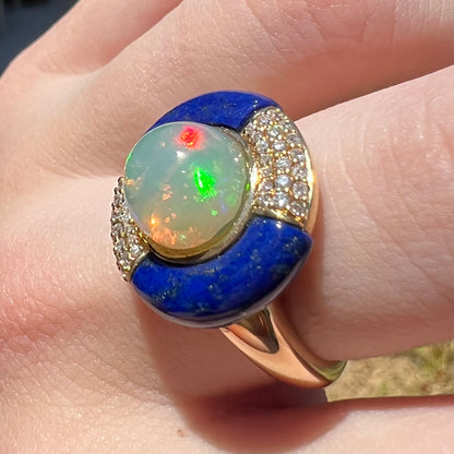 Angled close-up of a women's designer Ethiopian opal, lapis lazuli, and diamond ring in 14k yellow gold, shown on a finger in sunlight.  The opal has red and green flashes.