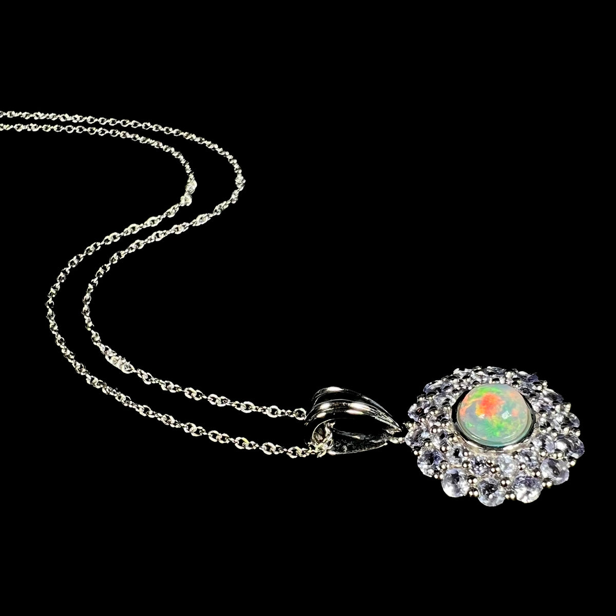 Angled view of a women's Ethiopian fire opal and tanzanite double-halo necklace laying on a black background, its silver chain flowing behind it.