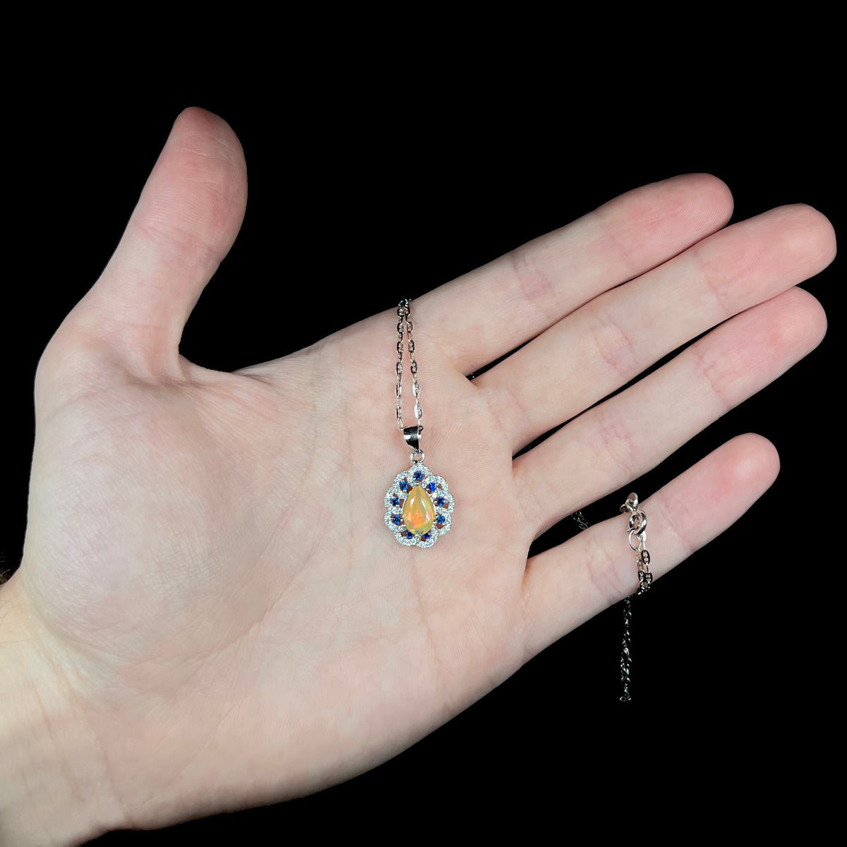 Women's Ethiopian opal, blue sapphire, and cubic zirconia necklace, held in hand; the chain is wrapped around pinky finger