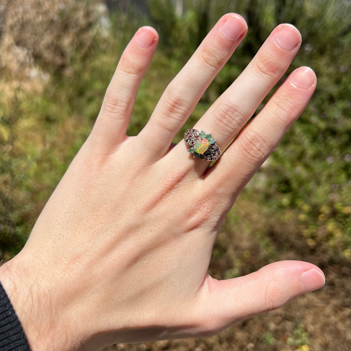 Hand wearing women's Ethiopian opal ring with garnet and emerald accents in natural sunlight