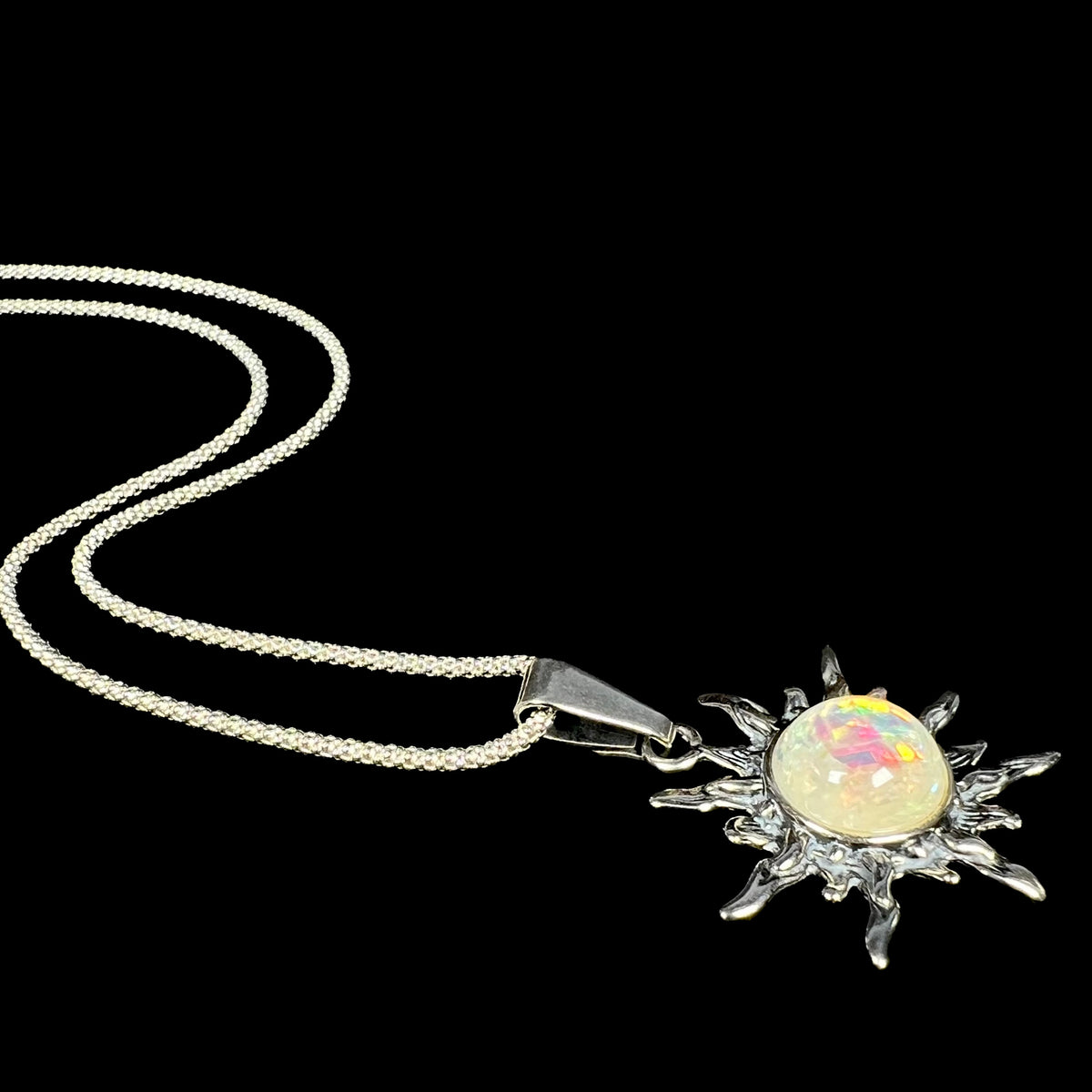 Angled side view of a women's faux opal sun necklace made from sterling silver, shown laying on a black background.  The pendant's chain flows behind it.