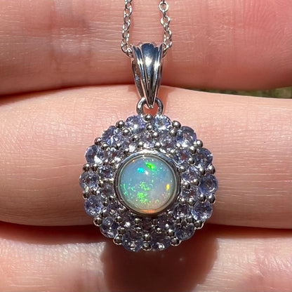 Close-up sunlit view of a women's Ethiopian fire opal and cool-blue double tanzanite halo pendant in sterling silver, resting on fingers.