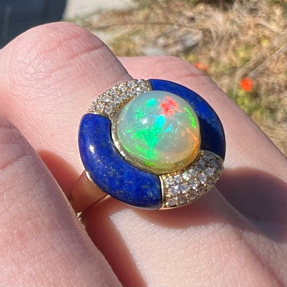 A close-up of a women's ring worn on a finger in sunlight, featuring a round iridescent fire opal center with green, blue, and red flashes, surrounded by deep blue lapis lazuli segments and small pavé-set diamodns on a 14k gold shank.
