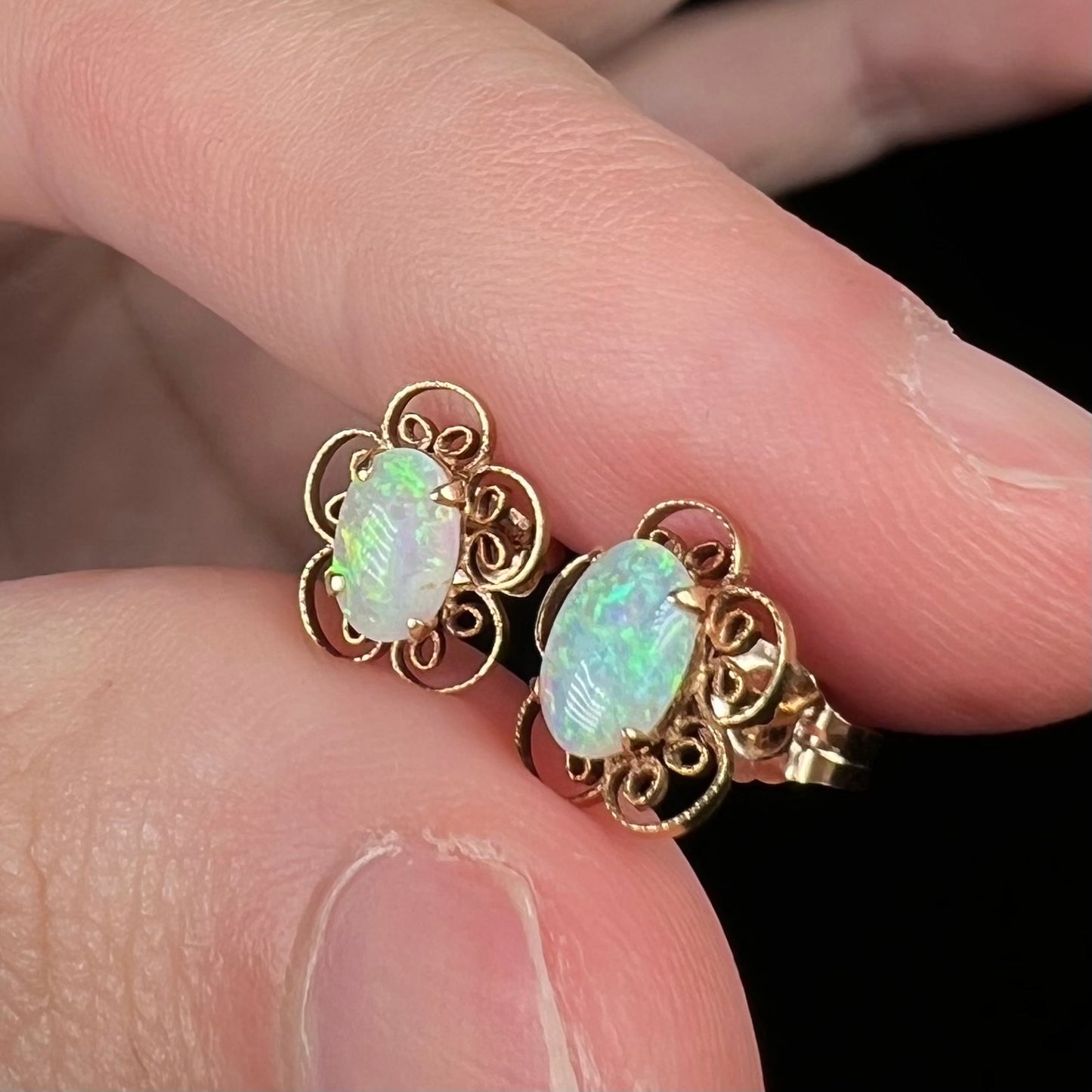 Women's 14k yellow gold opal stud earrings with filigree flower design, held between fingers
