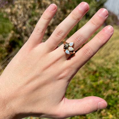Hand wearing a women's 14k gold three-stone Australian crystal opal ring in natural sunlight against blurred outdoor backdrop.