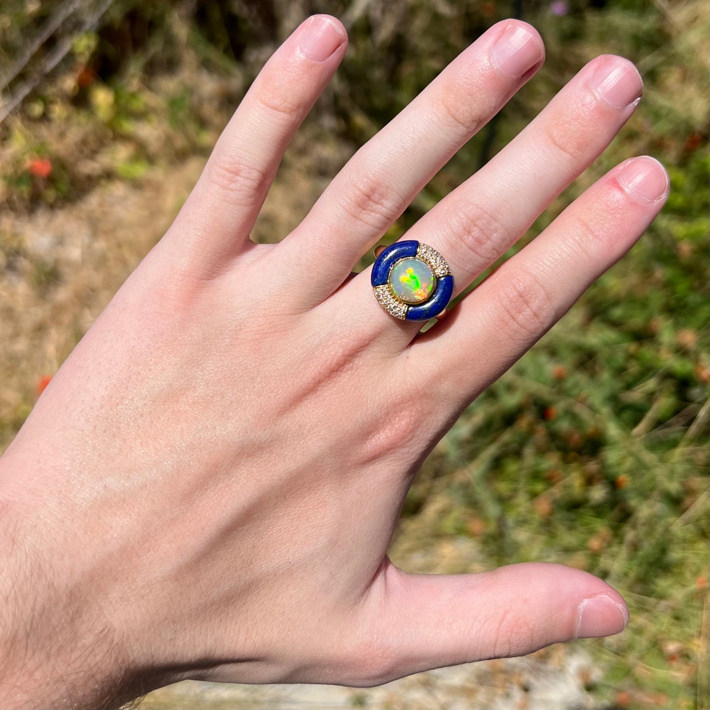 Hand wearing a large, round fire opal, lapis lazuli, and diamond women's ring made from 14k yellow gold in natural sunlight against a blurred green outdoor background.