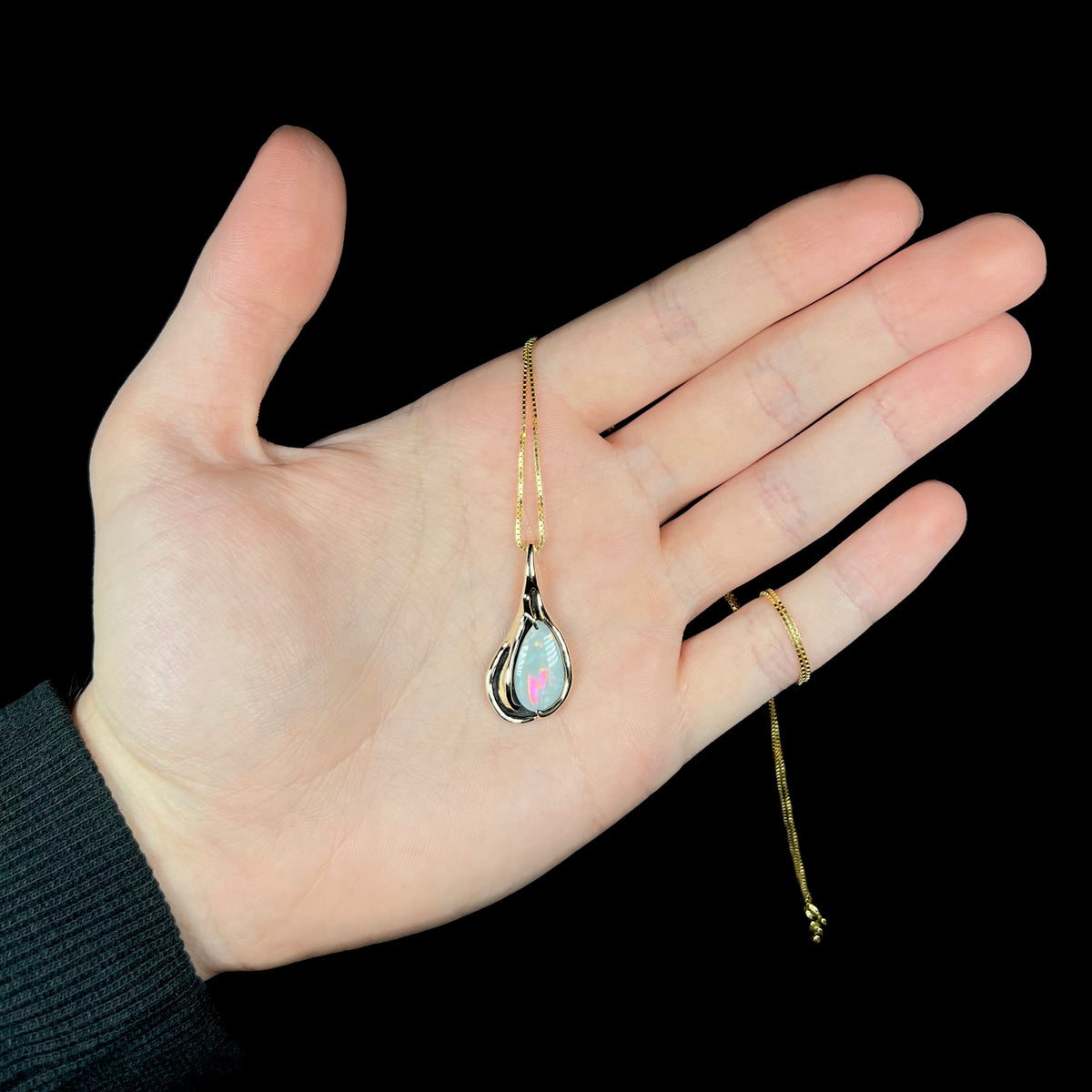 Hand holding women's Modernist style 14k gold opal necklace beneath LED studio light; the opal has a pinkish red flash of color.