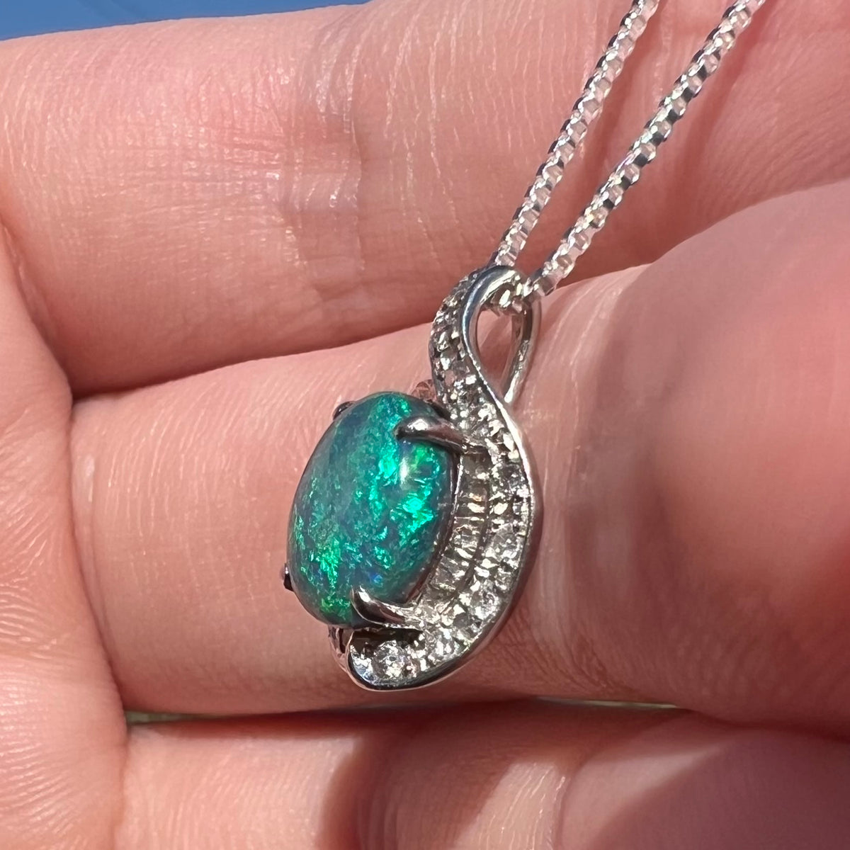Side close-up view of a natural, solid black opal women's pendant in platinum with diamond accents, held in a hand under sunlight.