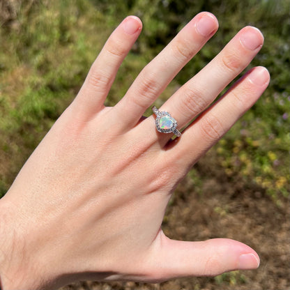 Hand wearing women's heart shaped opal and cubic zirconia halo ring in natural sunlight