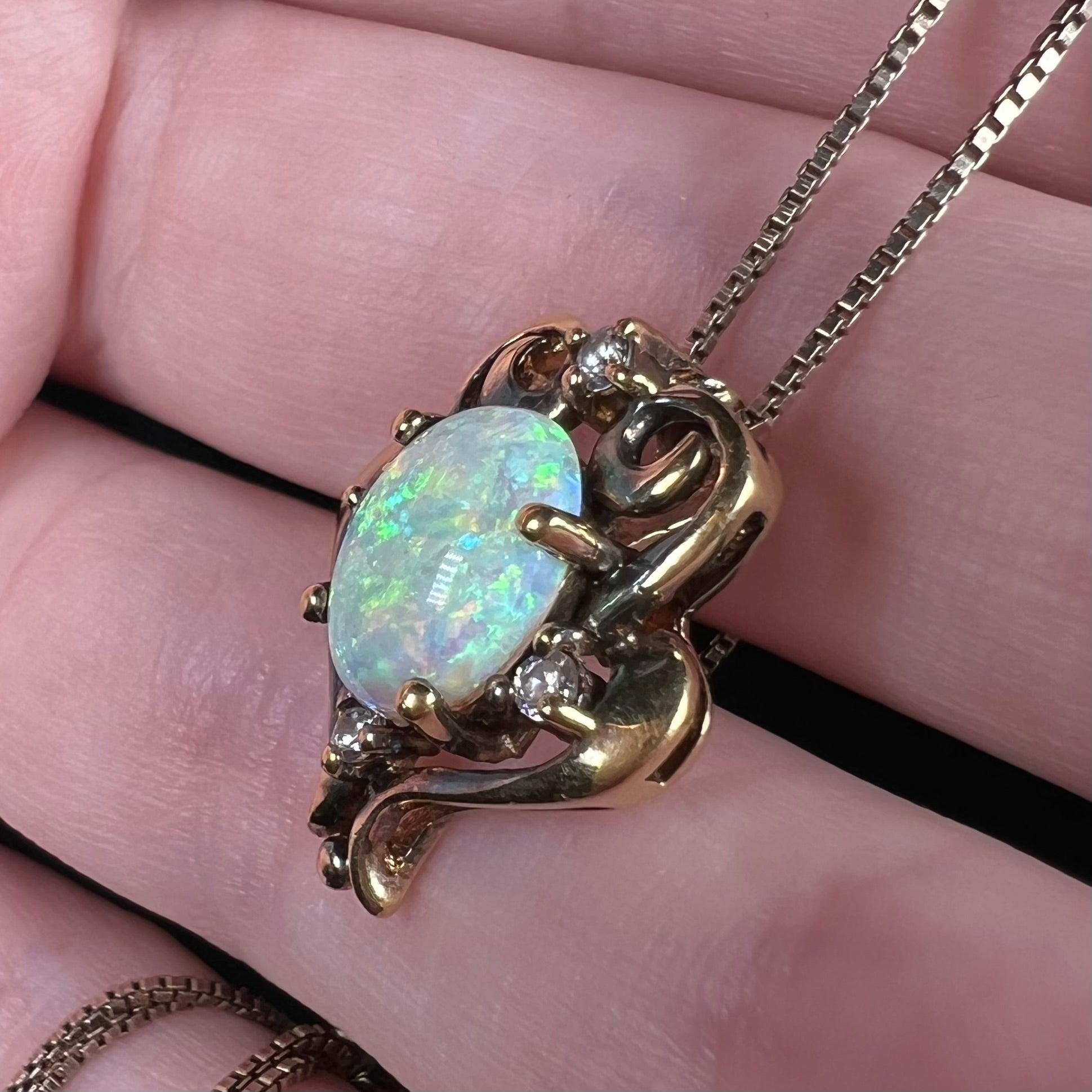 A women's synthetic opal and CZ necklace with a gold-tone Victorian style setting, shown close-up from an angle on fingers.