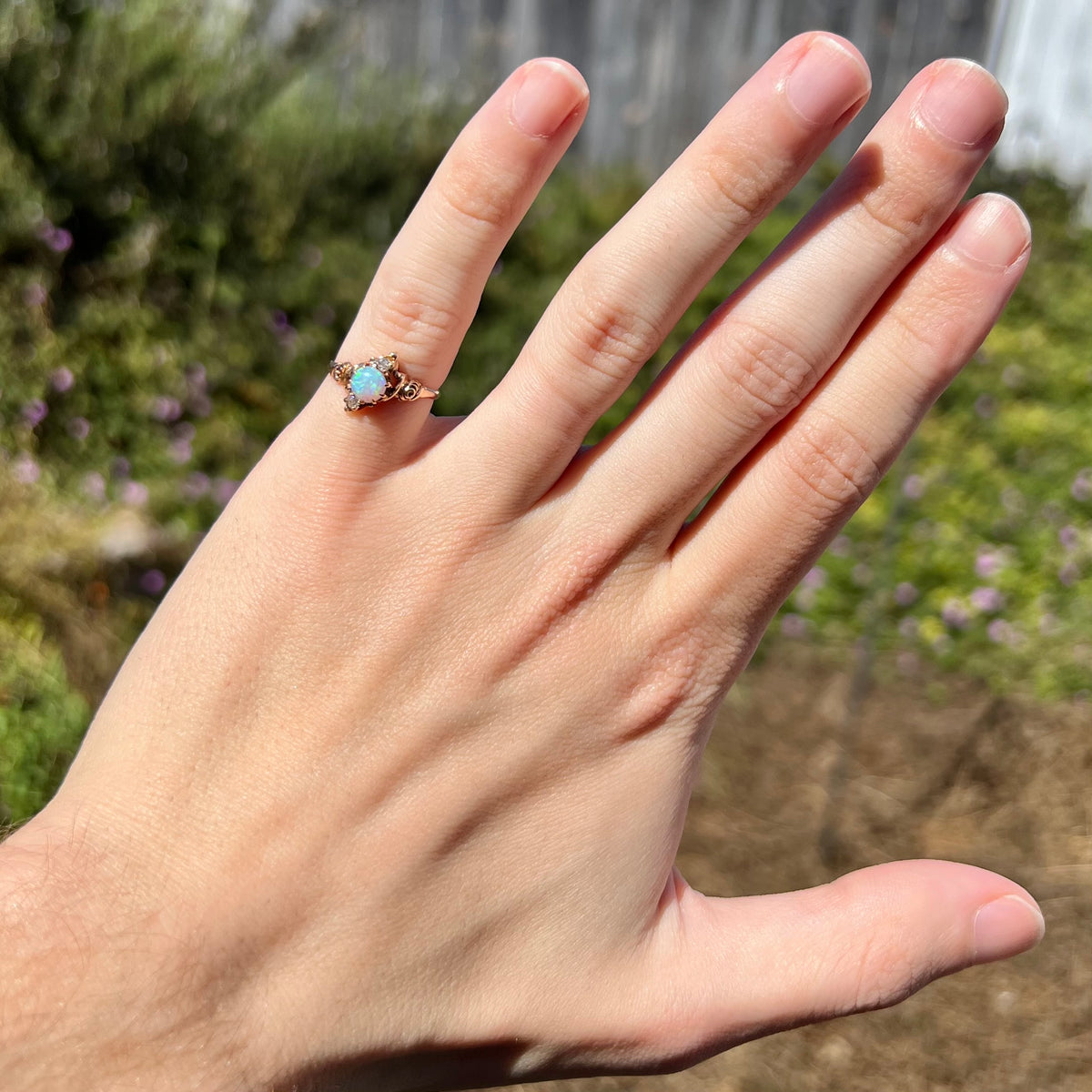 Sunlit hand wearing a women's gold Art Nouveau Australian opal and diamond pinky ring against blurred green outdoor background.