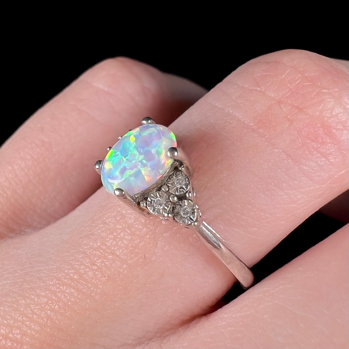 Macro view of women's sterling silver ring with lab-created opal center worn on finger, focusing on illusion-set diamonds in shoulder of ring.
