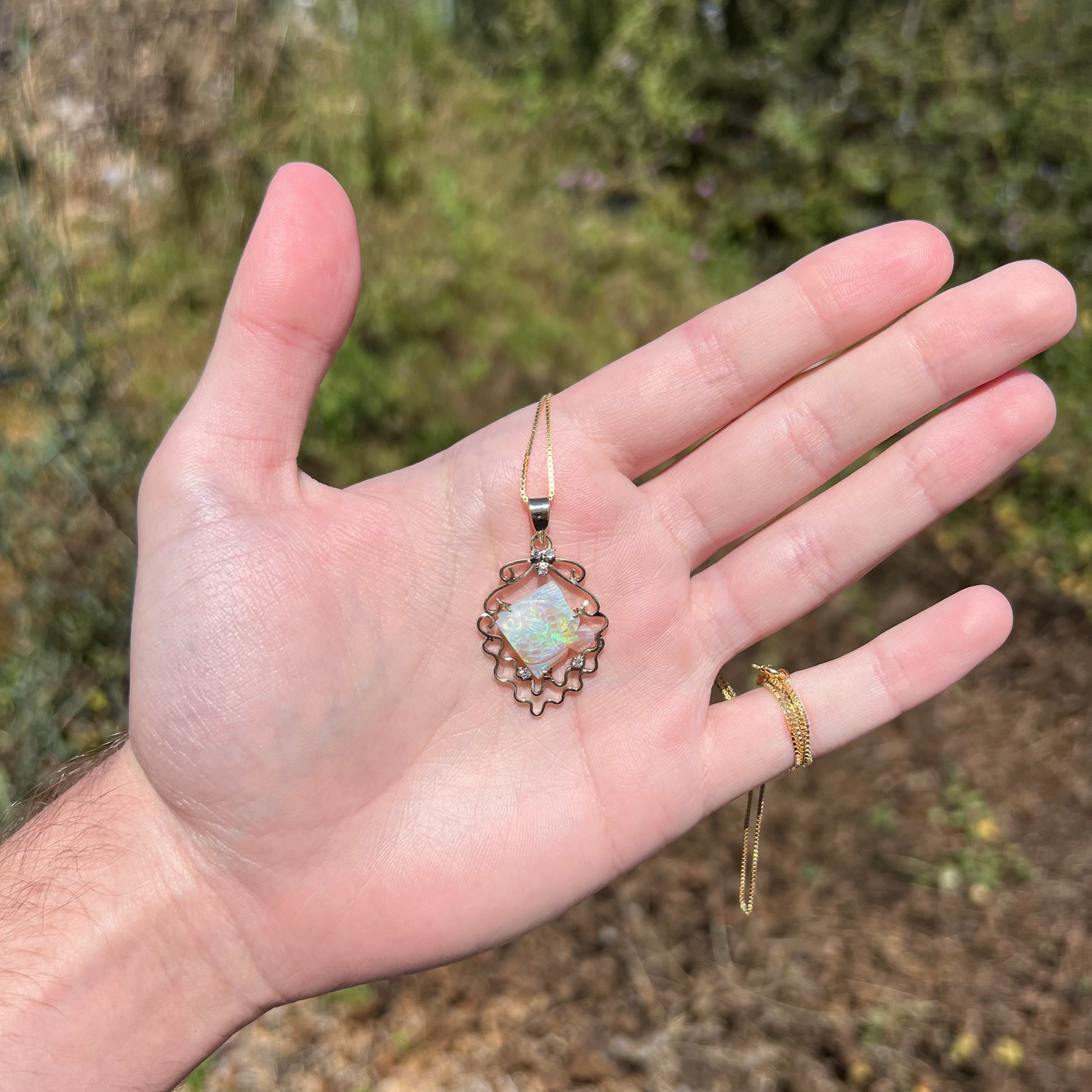 Hand holding women's carved Australian opal fish necklace with diamond accents in gold under sunlight