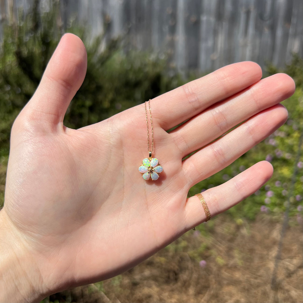Hand holding women's lab-created opal flower necklace in the sunlight; the pendant's 14k gold chain wraps around the hand's pinky finger