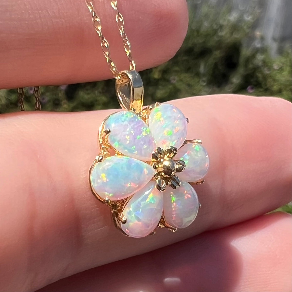 Angled close-up of a women's synthetic opal flower pendant set in 14k gold, shown held in a hand under natural light