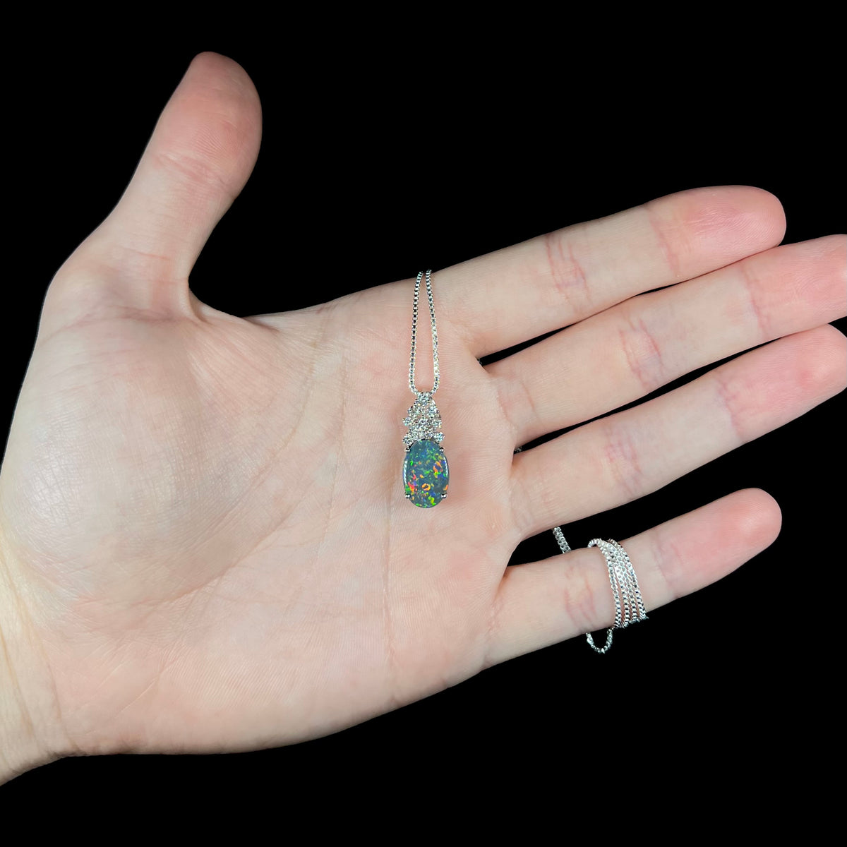 Hand holding women's black opal pineapple pendant in platinum with natural diamond crown on silver display chain, green and red flashes seen in opal.