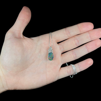 Hand holding women's black opal pineapple pendant in platinum with natural diamond crown on silver display chain, green and red flashes seen in opal.
