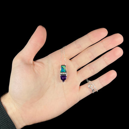 Hand holding a women's lab-created opal and trillion cut purple stone drop pendant beneath cool LED studio light.  The sterling silver chain is wrapped around the hand's pinky finger.