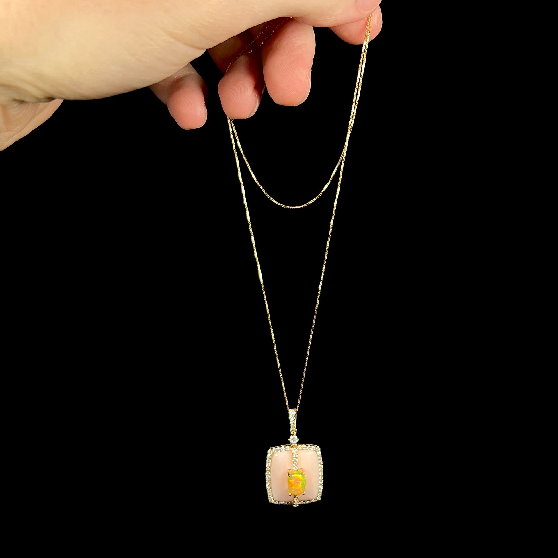Hand dangling a women's 14k gold pink opal and diamond necklace from its gold box-style chain.