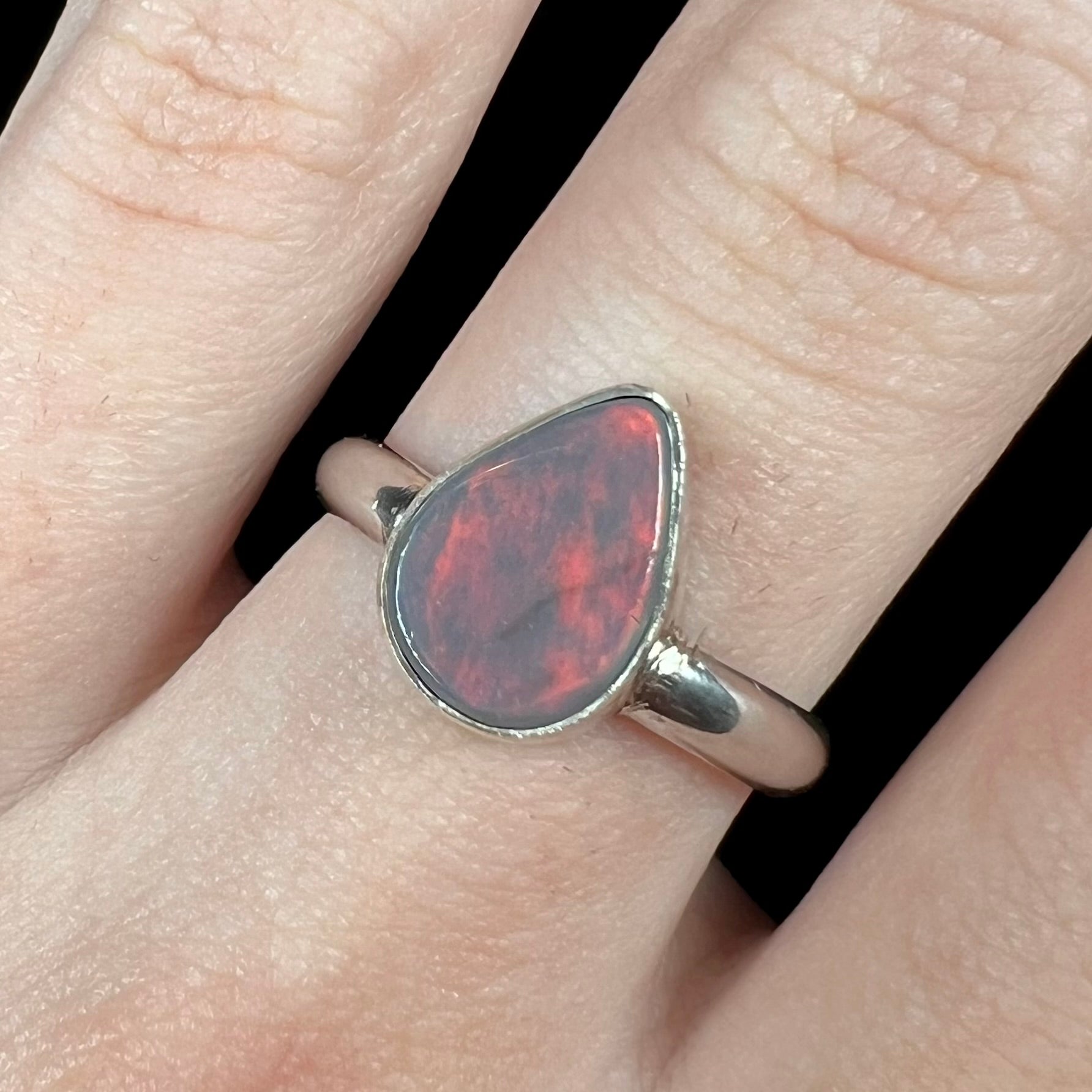 Close-up of a women's Australian opal solitaire ring in sterling silver, worn on a finger.  The pear shaped opal has a soft red flash of color.