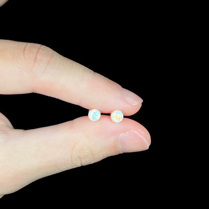 Fingers holding a pair of women's round lab-created opal stud earrings, mounted in 14k yellow gold prongs.