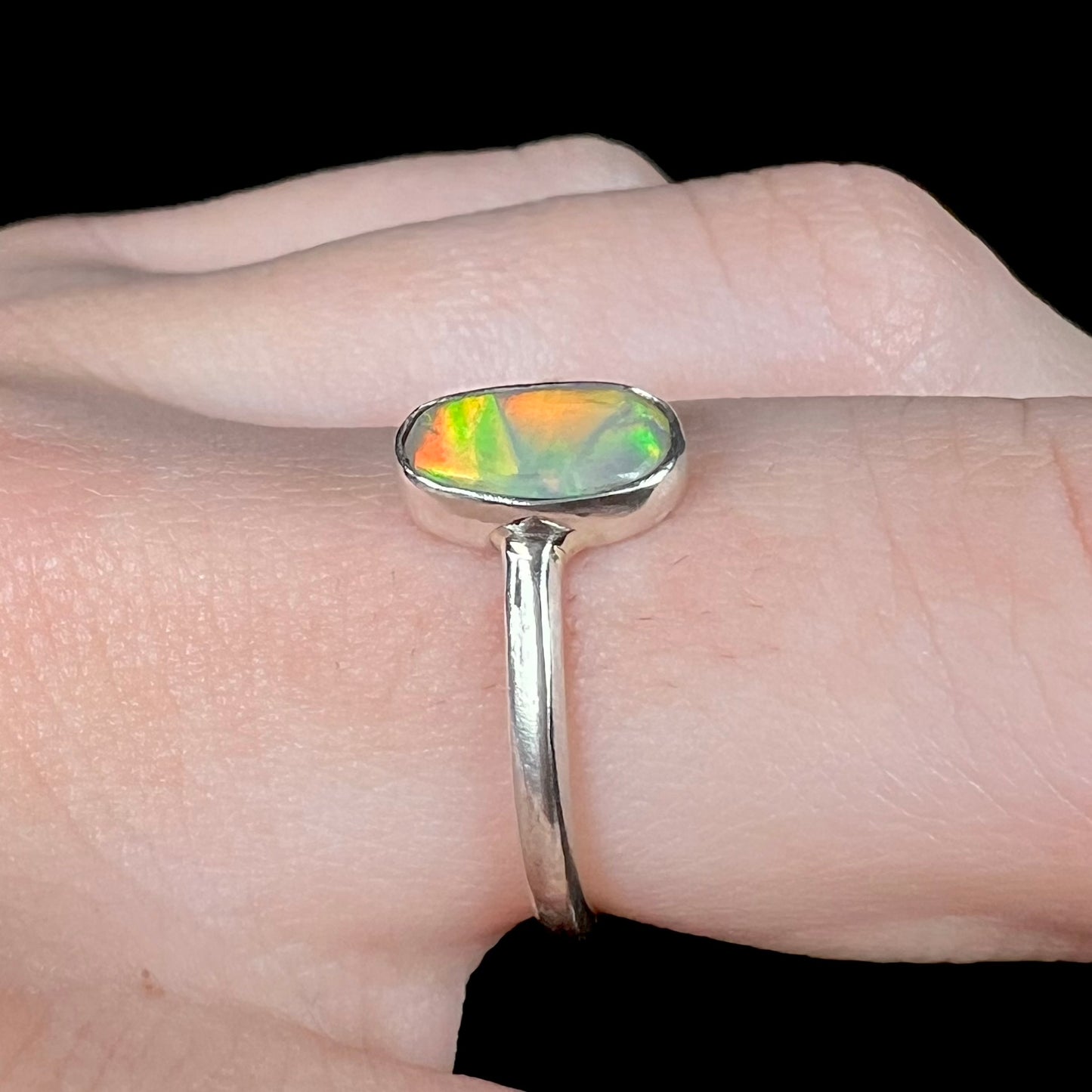 Side close-up view of a women's sterling silver Australian crystal opal ring, worn on a finger.  The opal has orange and green flashes.