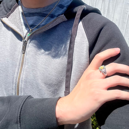 A close-up of a person wearing a black and gray zip-up hoodie, showing a ring on their finger. The ring features a silver-toned band with a large, colorful gemstone cluster.