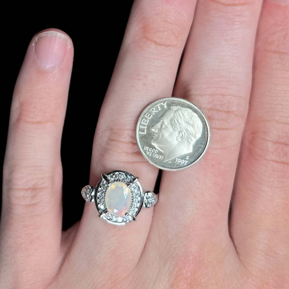 Hand wearing women's sterling silver Ethiopian opal and cubic zirconia halo ring next to US dime coin for scale