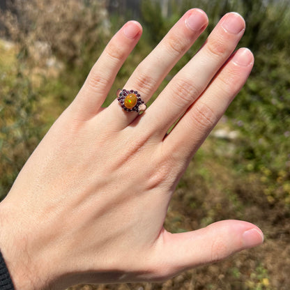Hand wearing women's sterling silver Ethiopian fire opal ring with bowtie-shaped garnet halo design in natural sunlight