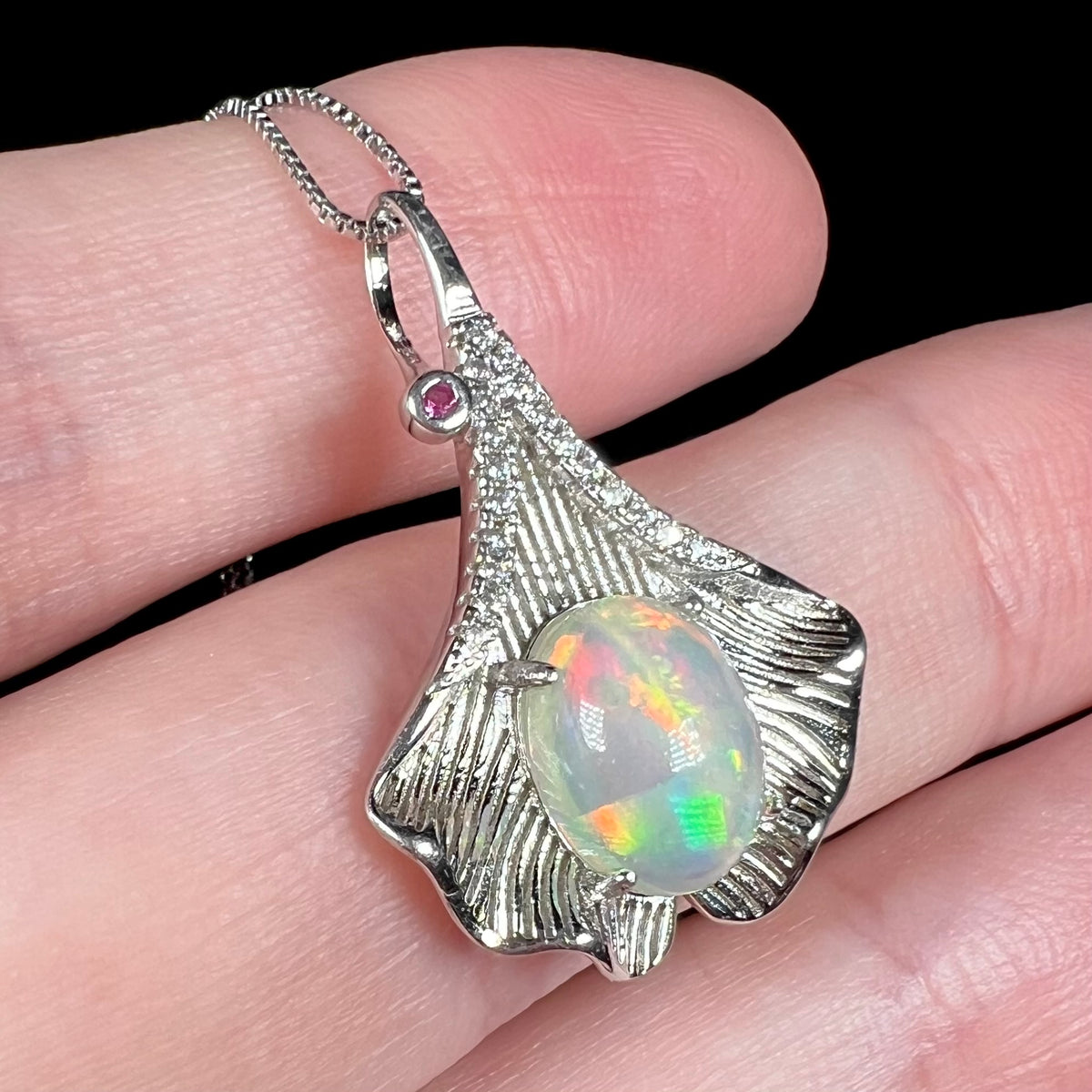 Women's sterling silver ginko leaf pendant with Ethiopian opal, pink sapphire, and CZs, held in hand
