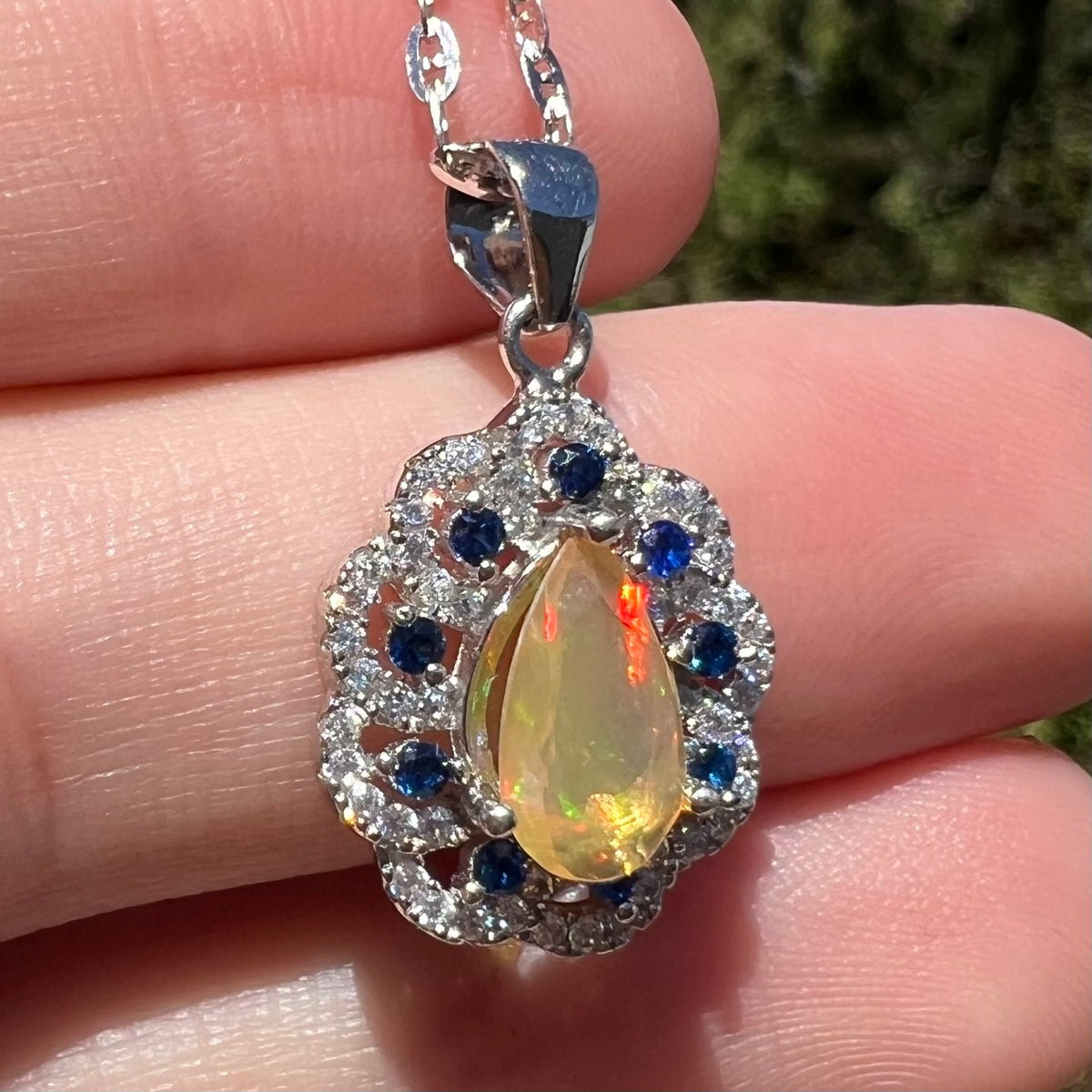 Women's sterling silver Ethiopian opal pendant with sparkling CZ and blue sapphire accents, shown close-up in hand under natural sunlight