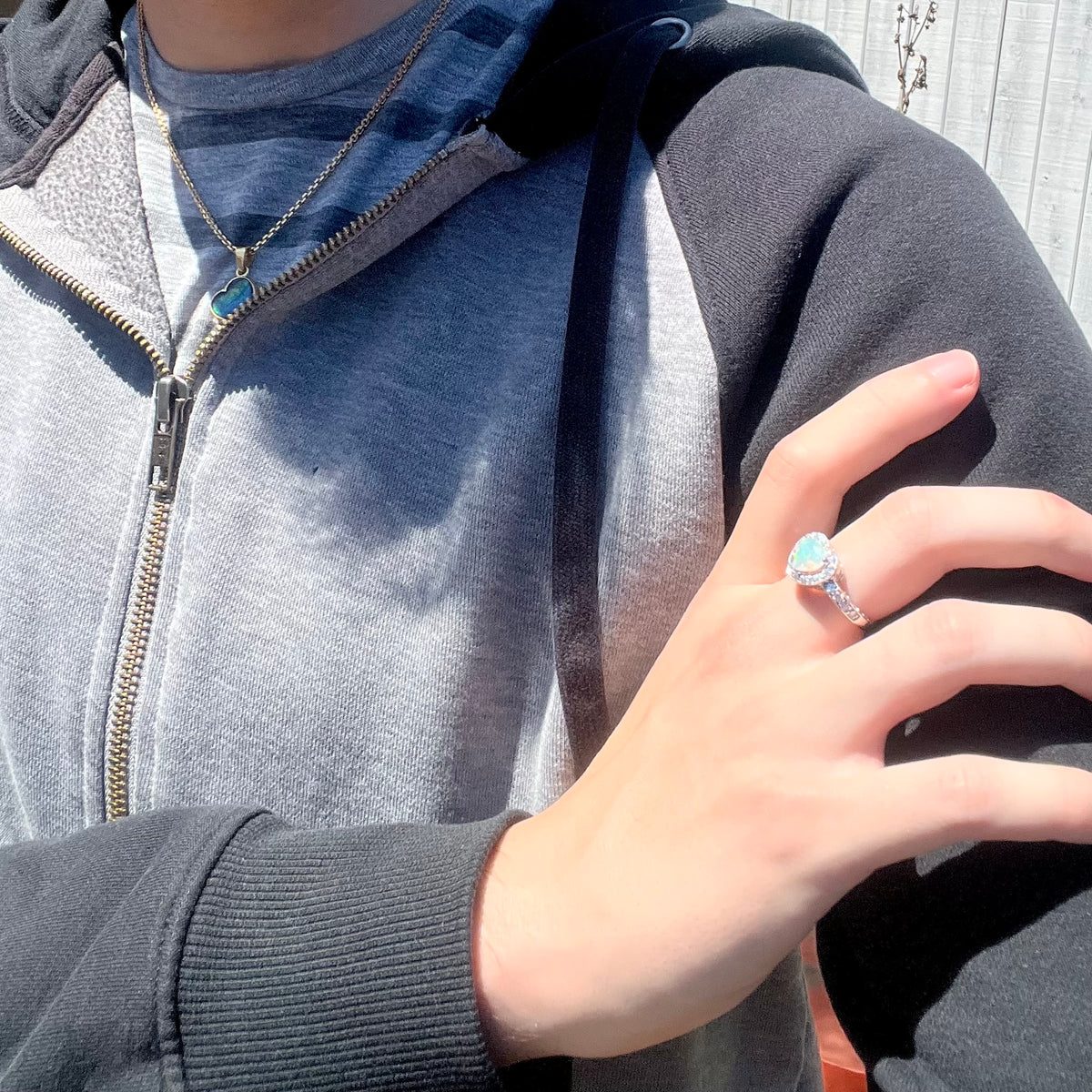 Close-up of person resting hand on opposing shoulder wearing women's silver heart shaped opal and CZ halo ring in sunlight