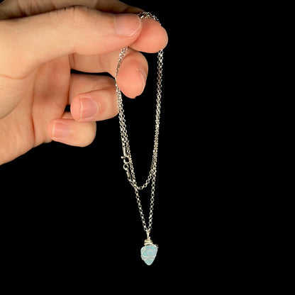 Hand dangling a women's silver wire wrapped opal doublet necklace from its base metal chain against black background.