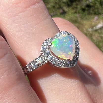Close-up sunlit angled view of women's sterling silver opal heart ring with cubic zirconia accents, worn on finger