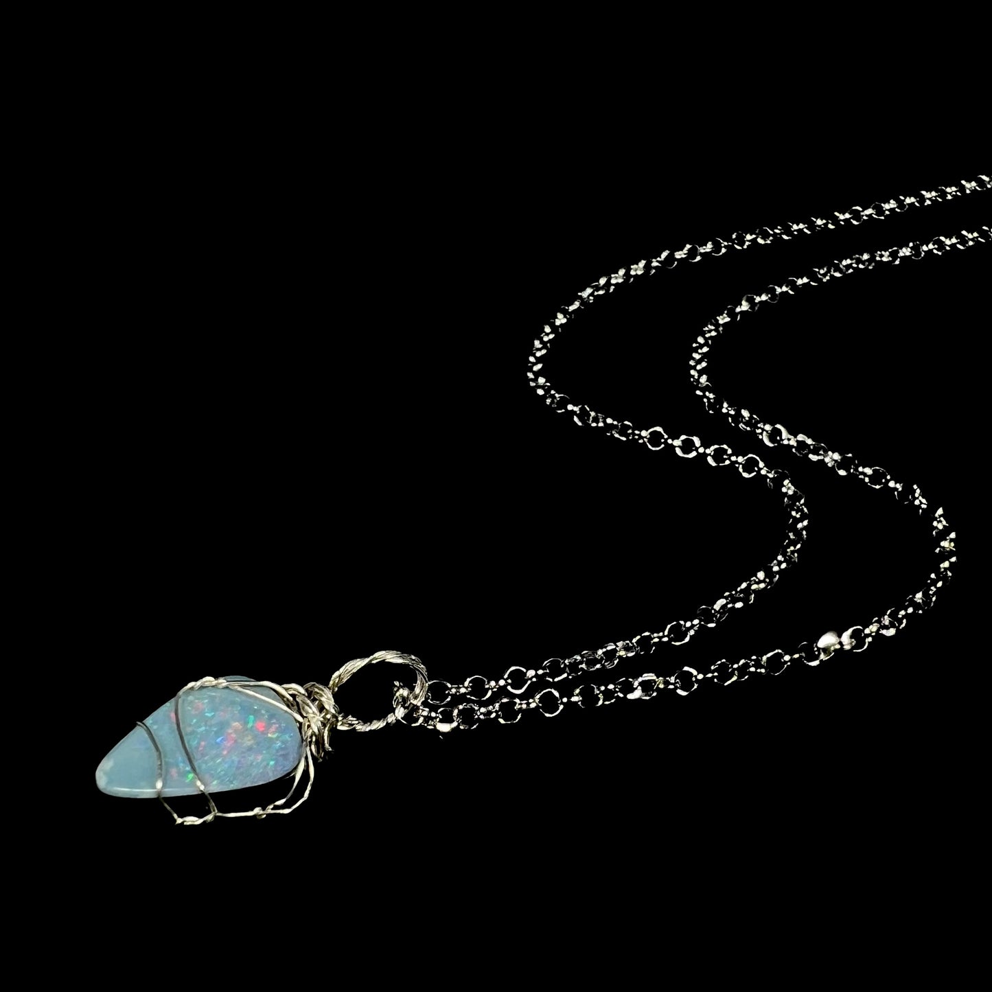 A women's freeform-shaped sterling silver wire wrapped opal doublet necklace shown from an angle laying on a black background, its chain flowing.